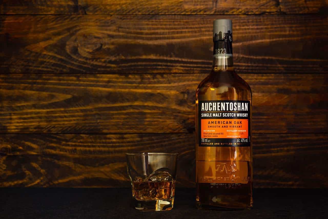 Bottle of Auchentoshan American Oak Scotch Whiskey and glass with ice on a dark wooden background. Triple-distilled single malt scottish whiskey.