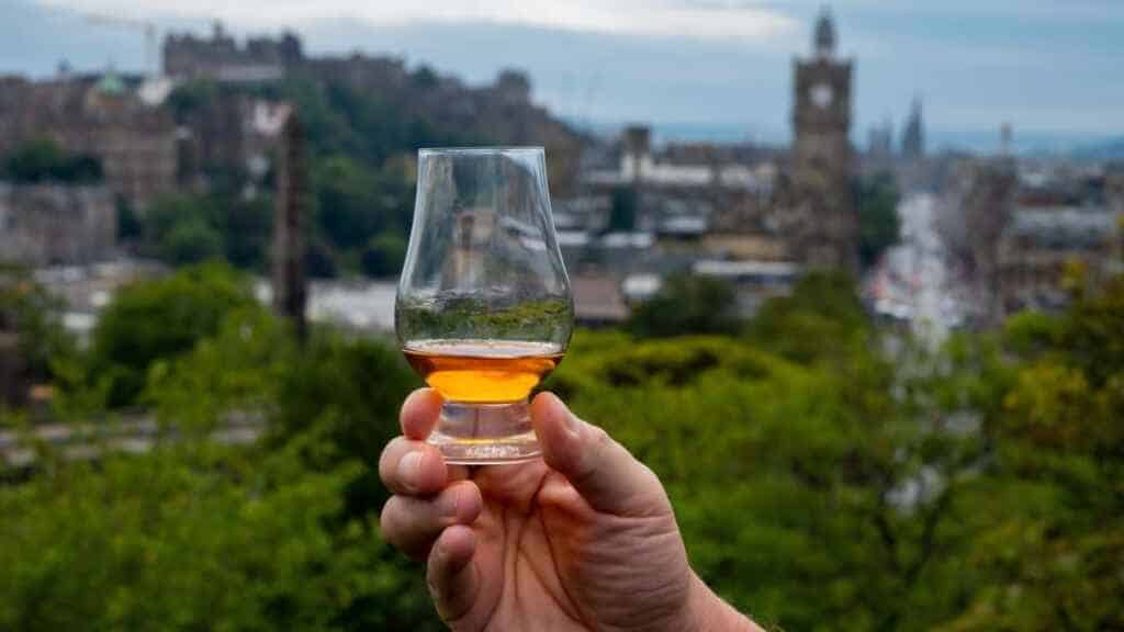 Hand holding glass of single malt scotch whisky and view from Calton hill to park and old parts of Edinburgh city in rainy summer day, Scotland, UK