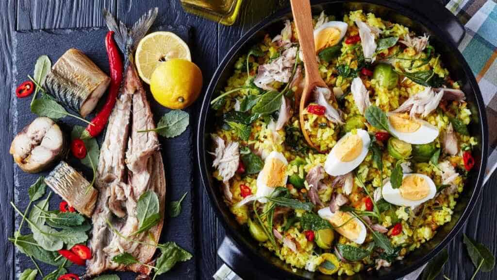 Delicious Kedgeree with flaked smoked fish, hard boiled eggs, rice, kale, brussel sprouts, spices and herbs in a dutch oven on a black wooden table with ingredients, view from above,