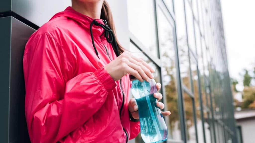 Woman in a rain jacket leaning against a building holding a reusable water bottle.