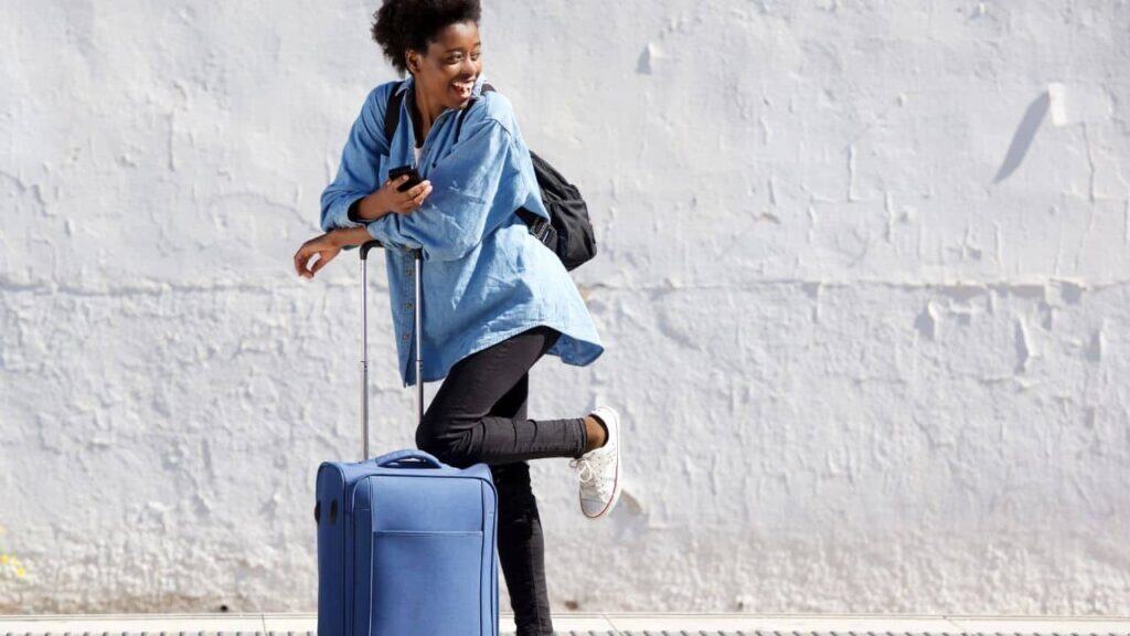 A woman in comfortable clothing leans on her roller suitcase outside a train station