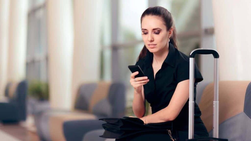 Young woman with a suitcase sitting at the airport on her phone.