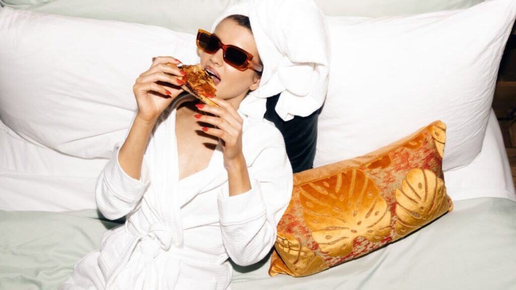 A woman wearing a robe with her hair up in a towel in a hotel bed eating a piece of pizza.