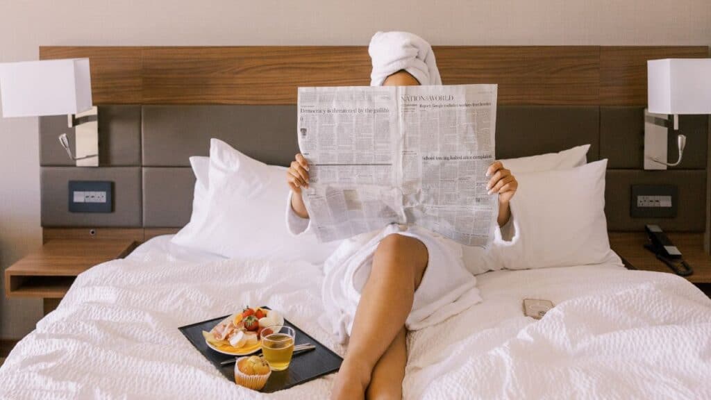 A woman wearing a robe with her hair up in a towel in a hotel bed reading the paper with breakfast in bed.